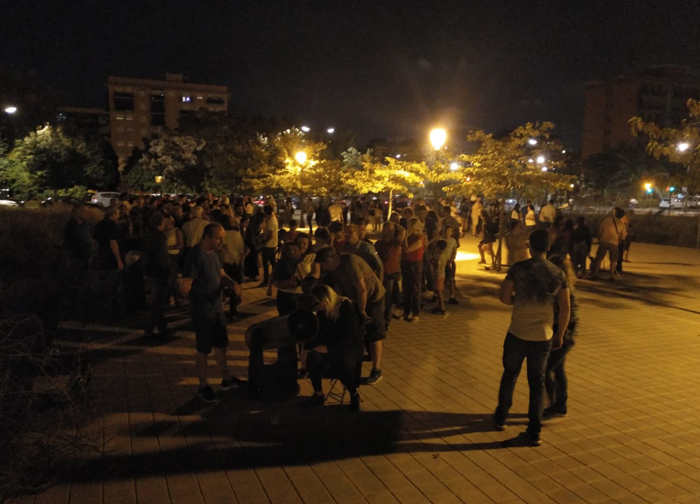 A small crowd observes the Moon at night, in a large paved area with trees in the background.