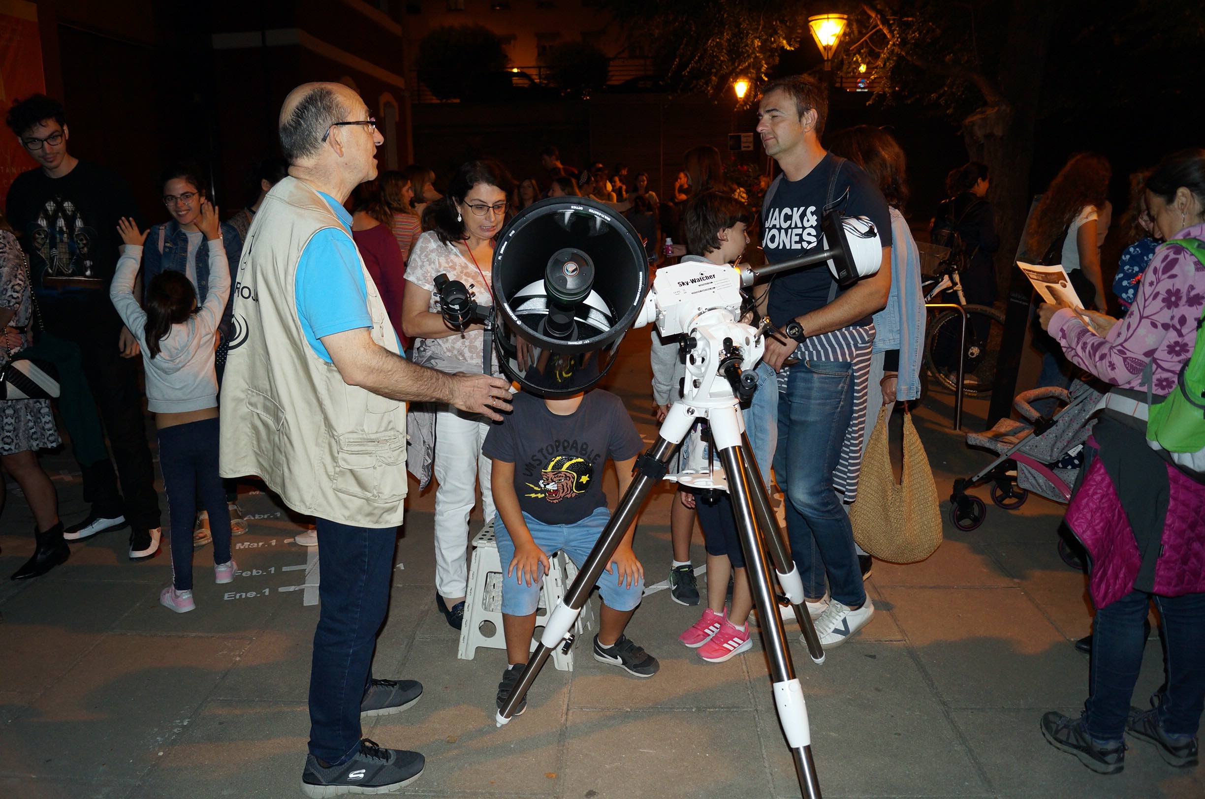 Two people have a conversation over a telescope, with other observers in the background. 