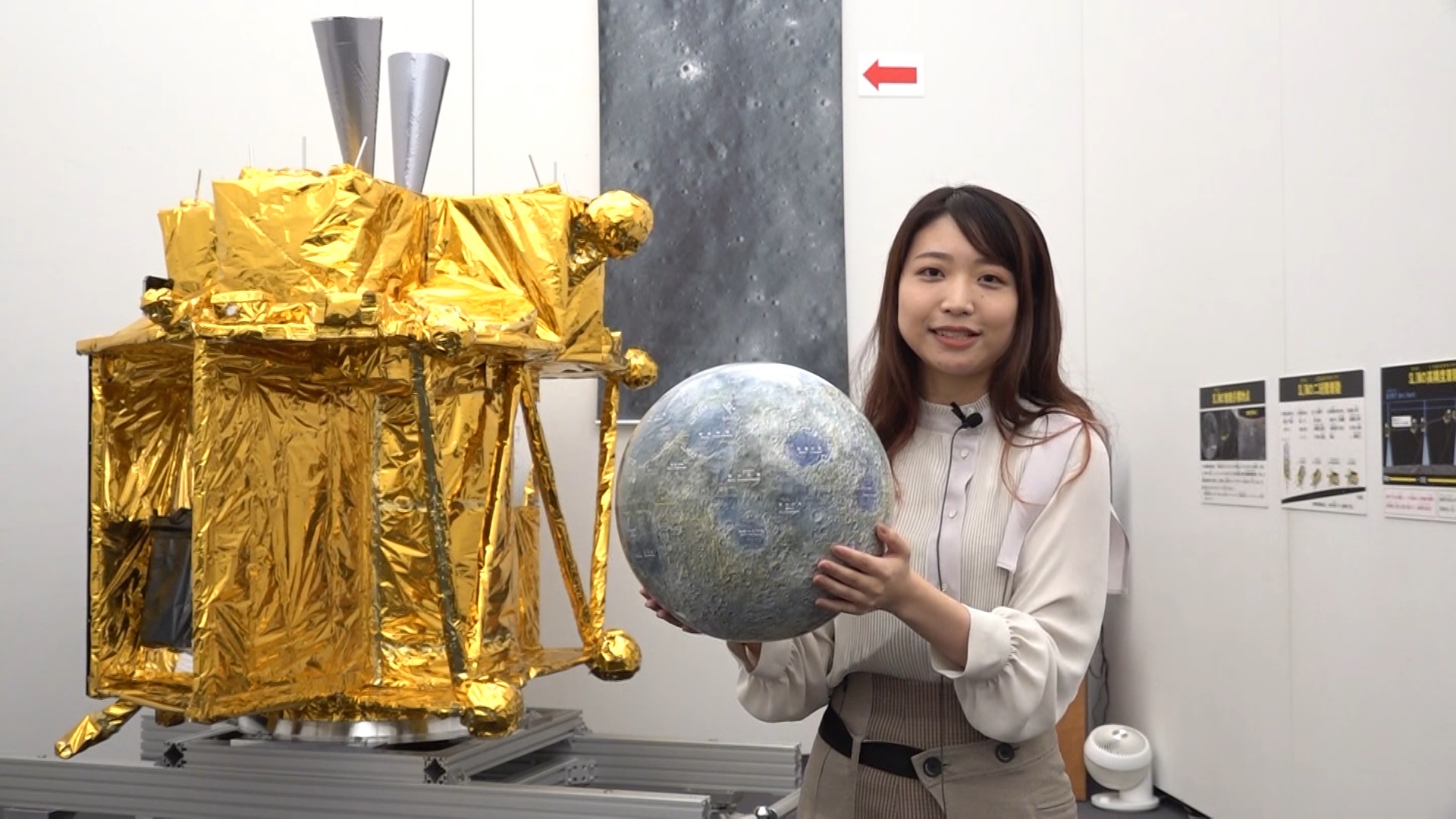 Photo of young woman holding spherical object in front of exhibit model of spacecraft in gold foil.