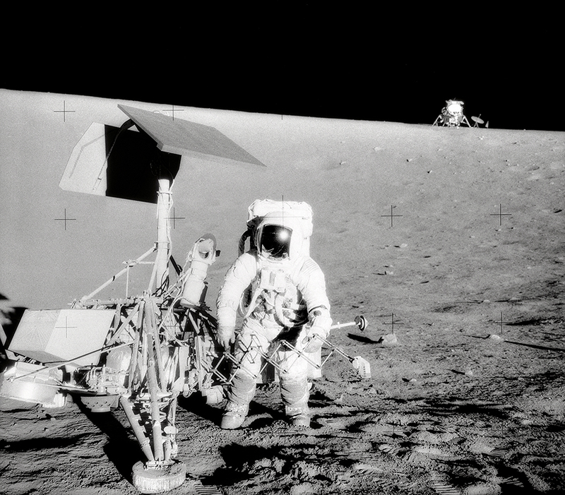 Black and white image of astronaut standing next to robotic spacecraft on the Moon.