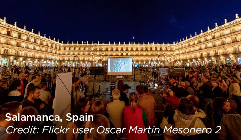 Large crowd of people in a town square attending a presentation event at with the Moon in the night sky