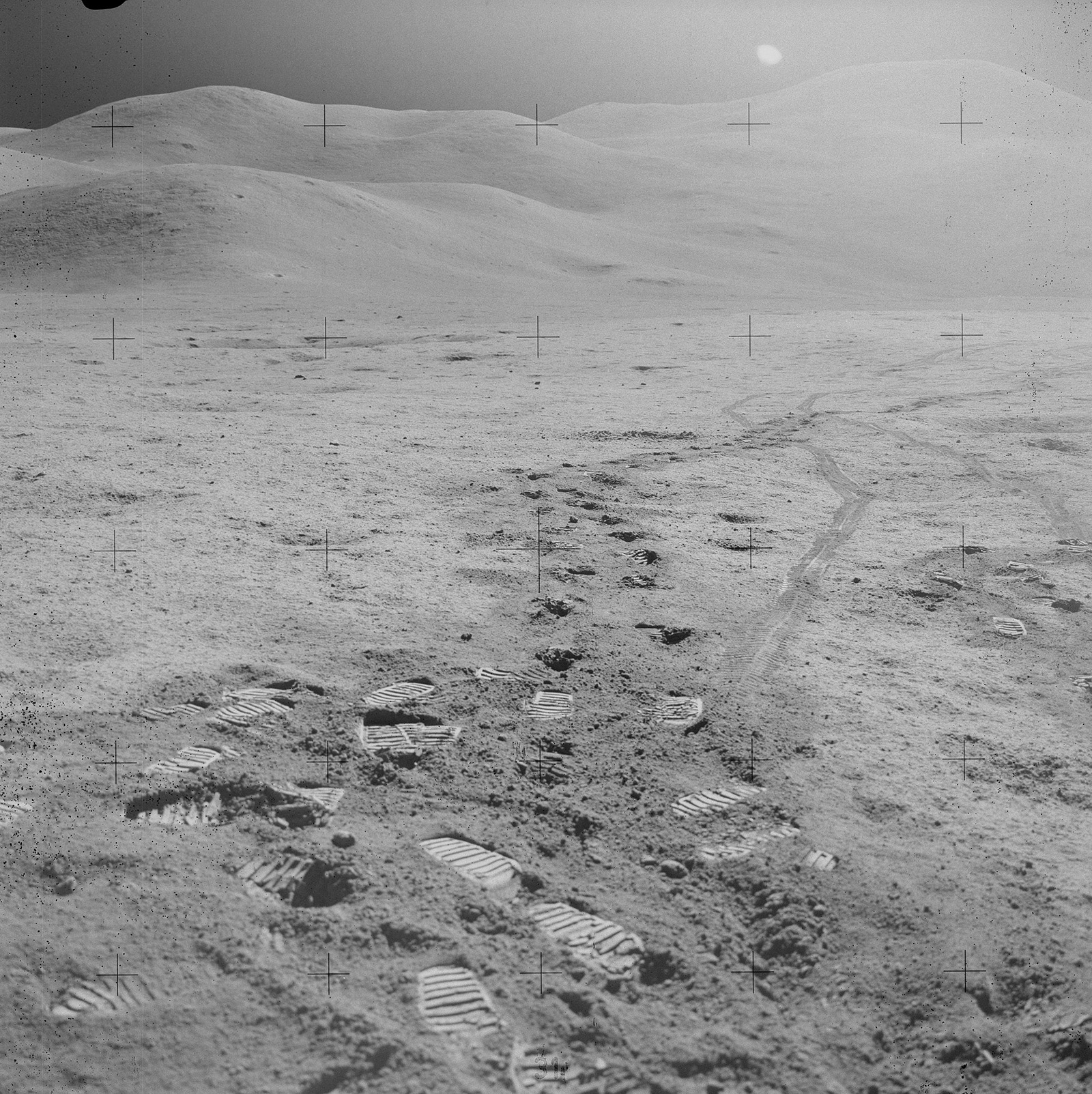 Grayscale image of a barren landscape. The Sun is visible in a dark sky. In the foreground, scattered bootprints show where astronauts have worked. More bootprints and rover wheel tracks lead into the distance, towards a lumpy mountain range. The lunar surface appears to have a fine, powdery texture, with some rocks.