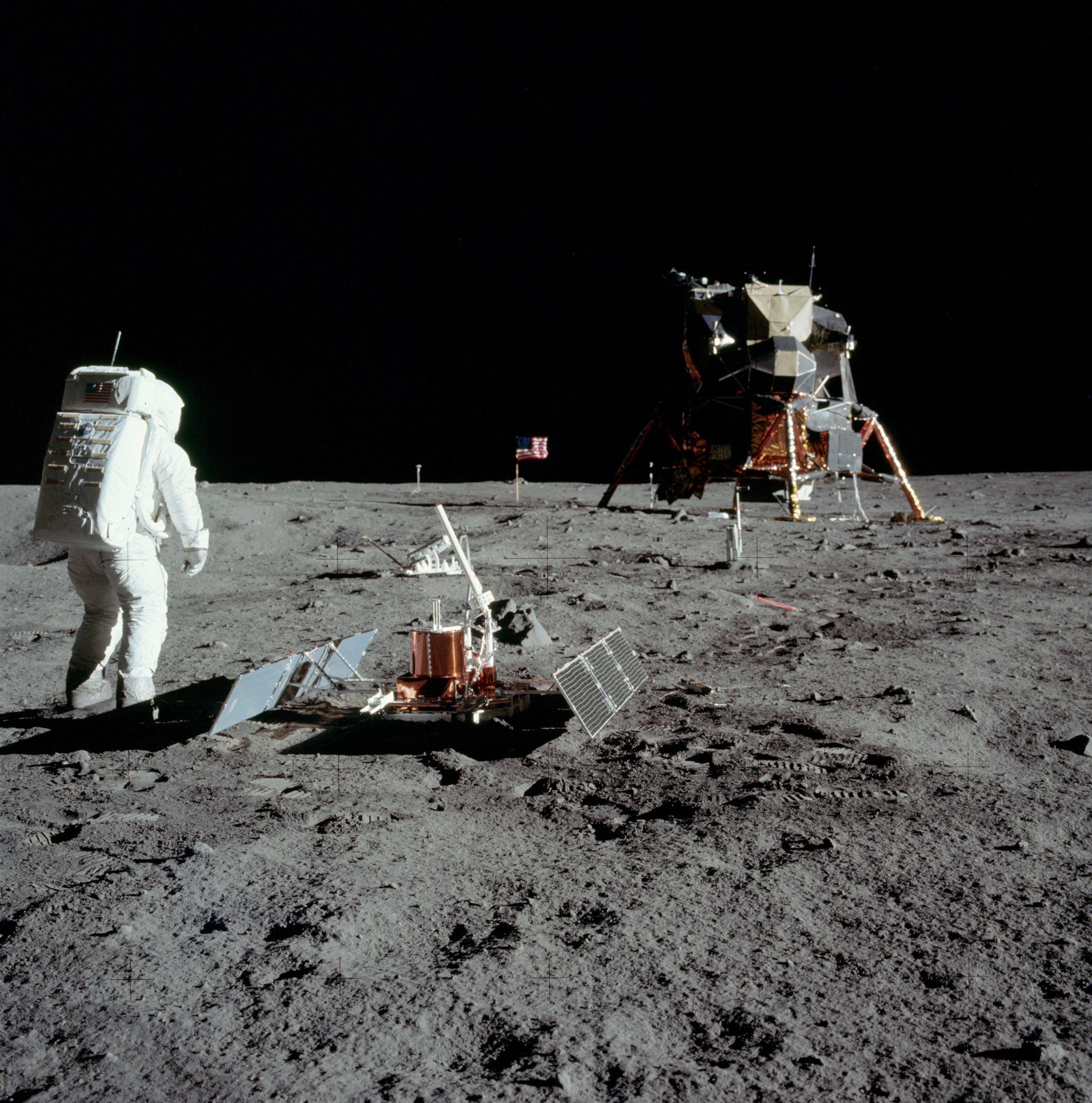 An astronaut stands on the Moon. He is in a spacesuit and his back is facing the camera. On his right is a copper-colored metal drum with two solar panel arrays on the left and right, extended like wings. A laser reflector is beyond the drum. Going backwards into the image, the lunar module stands on four legs and is copper and silver. Further back, an American flag is planted into the dusty Moon.