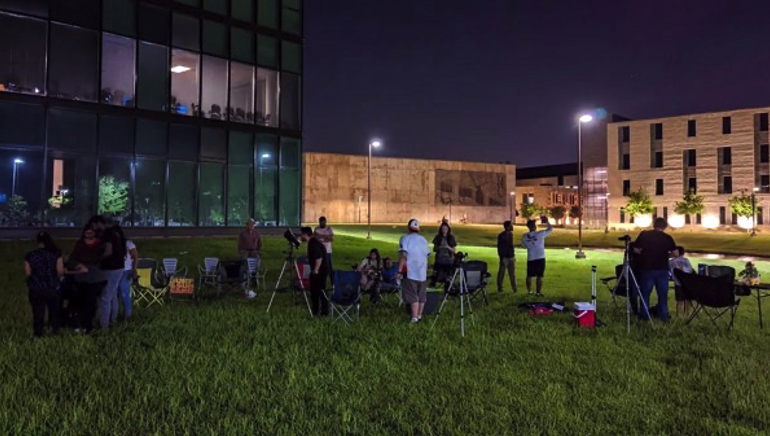 Photo of people gathered on a grass field at night. Lawn chairs and various cameras and telescopes are set out.