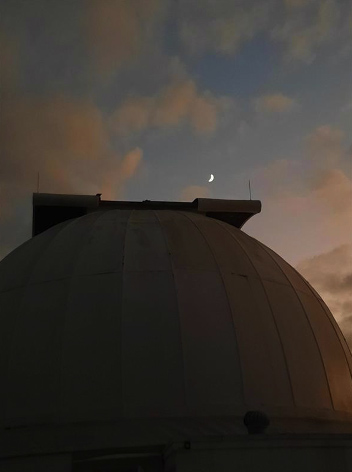 Photo of the evening sky with a tiny crescent Moon in the sky and clouds above a domed building structure.