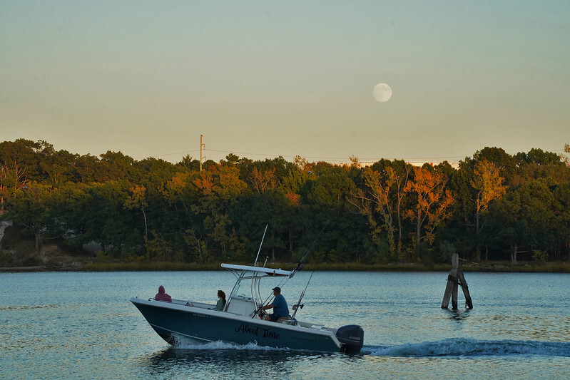 A small boat with three people aboard makes its way through the water. Trees that have begun changing color for autumn line the water in the background. A waxing gibbous moon rises above the tree line.