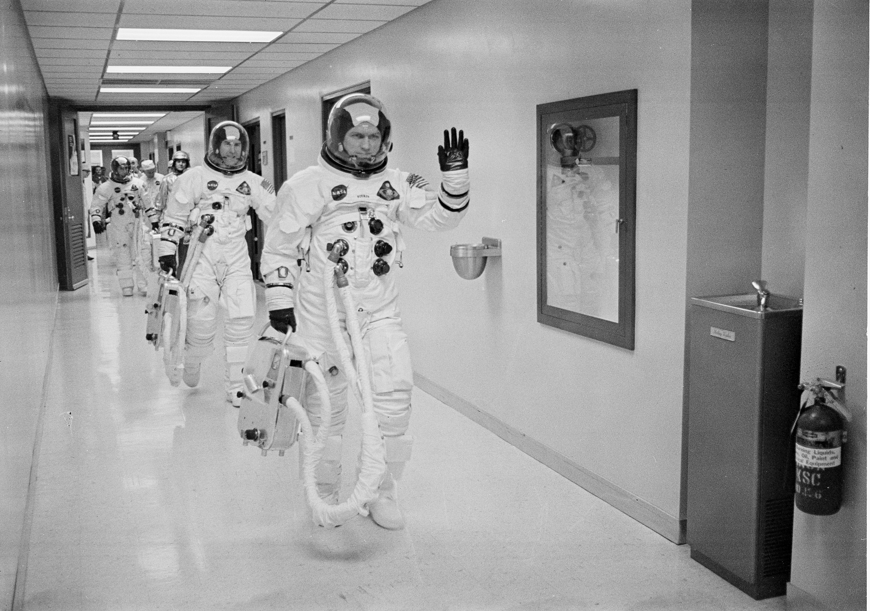 Trio of men in spacesuits walking down a hall.
