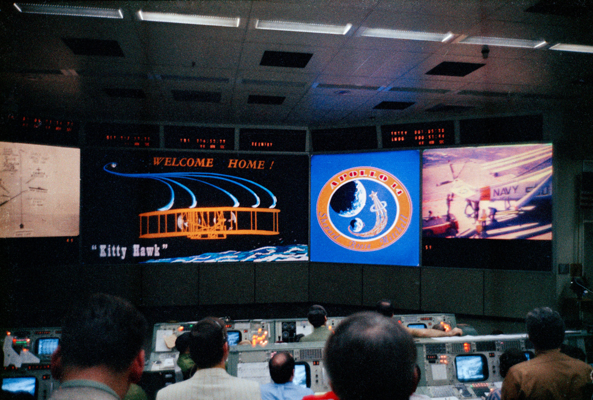 Mission control display monitors show "Welcome Home" message