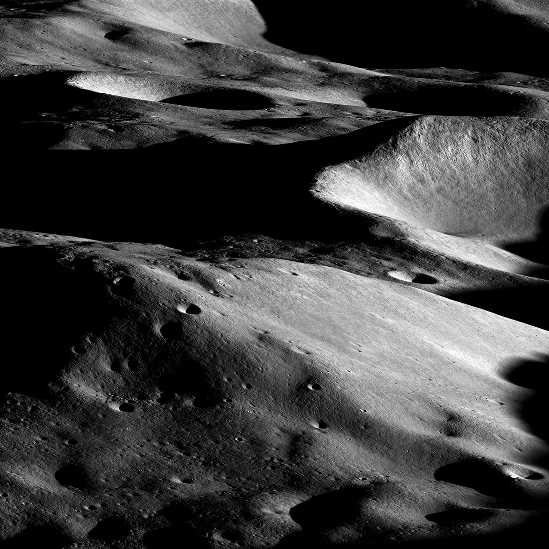 Dramatic grayscale image of a mountainous lunar landscape. Large craters with black shadows inside are visible in the background. In the foreground, a mountain is sunlit from the right and pocked with small craters. 