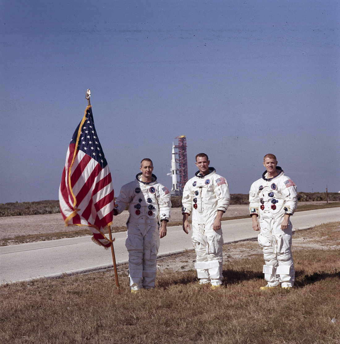 Three astronauts, one holding American flag, with Apollo 9 spacecraft in background