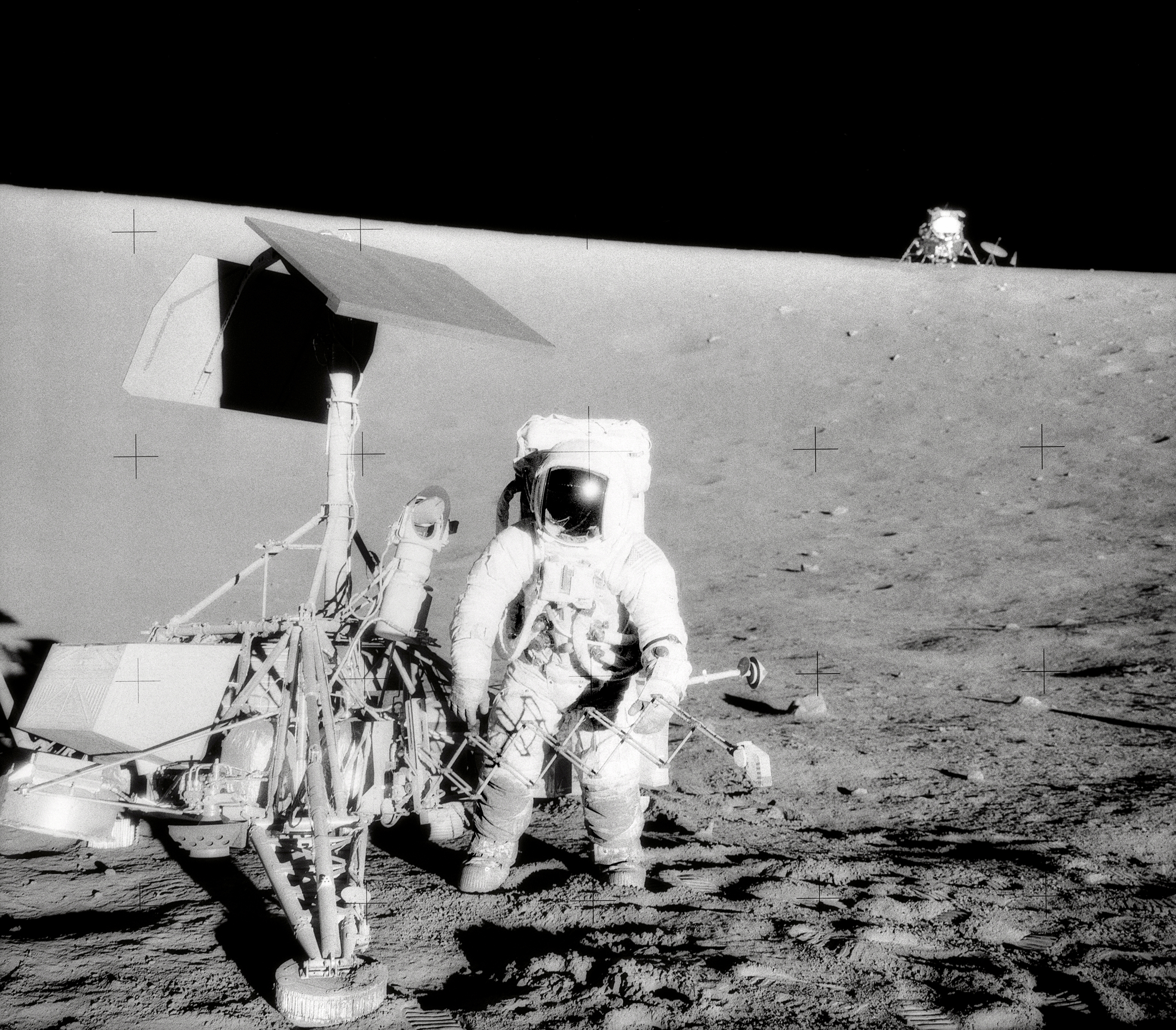 Black and white image of astronaut standing next to robotic lander on the surface of the Moon.