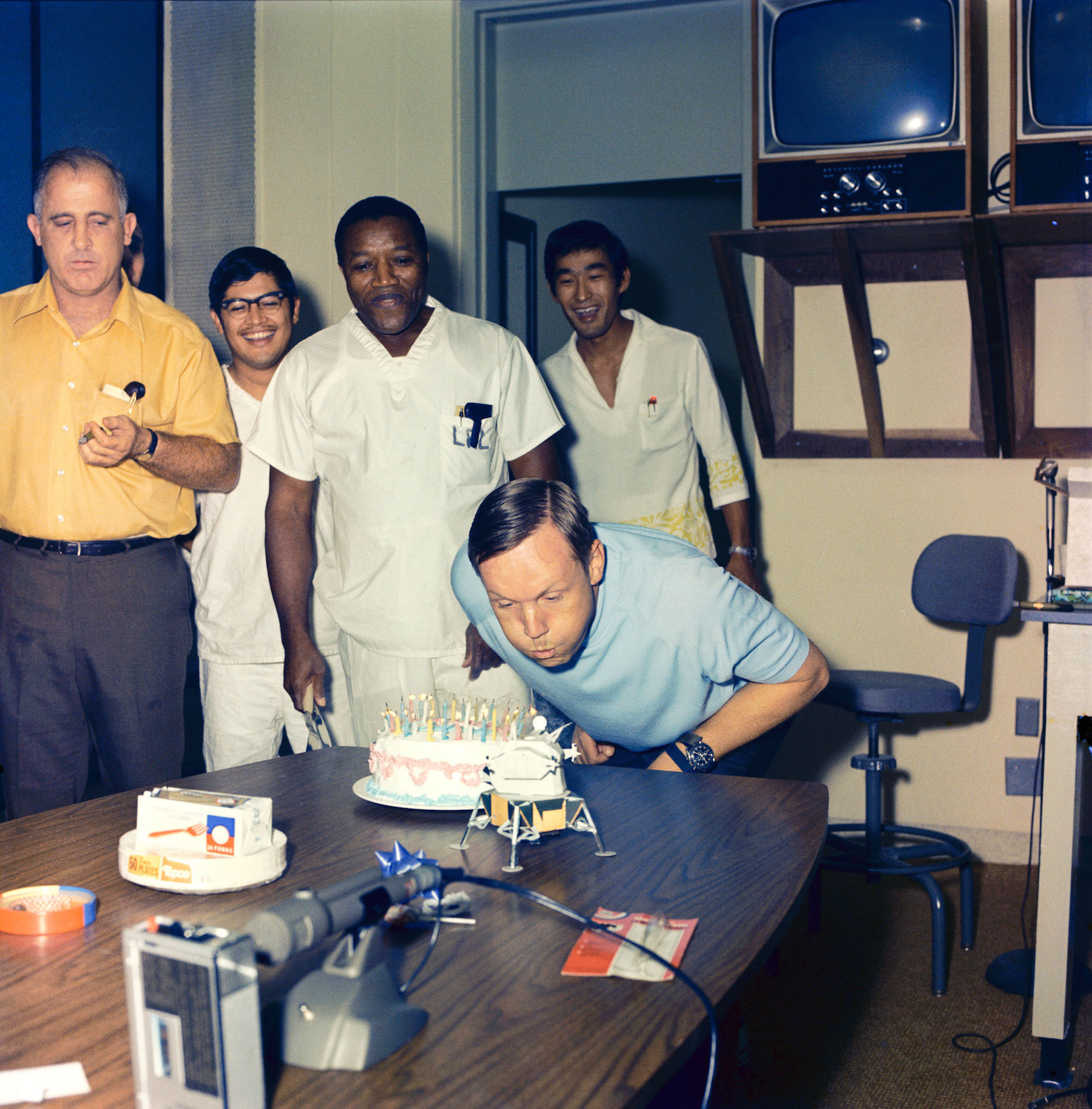 man blowing out birthday cake candles with people watching from behind