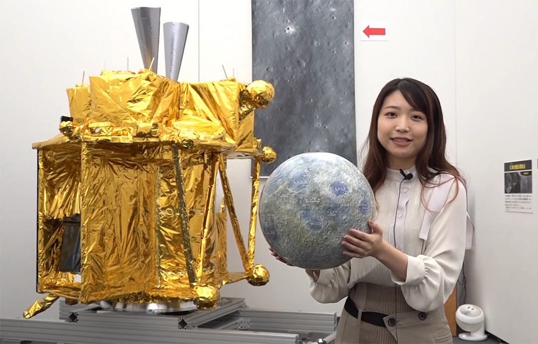 Photo of young woman holding spherical object in front of exhibit model of spacecraft in gold foil.