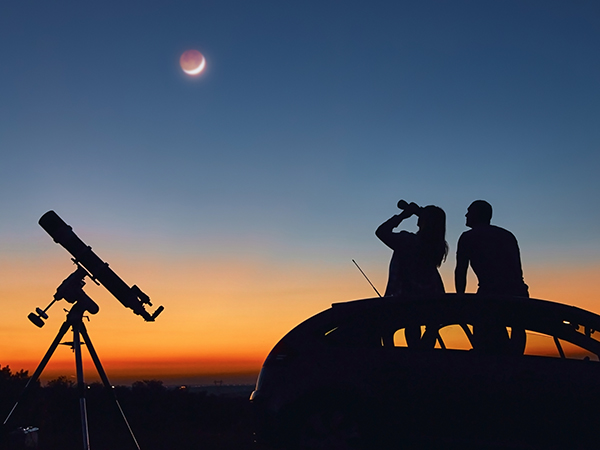 Silhouette of two people looking at the Moon in the sunset sky.