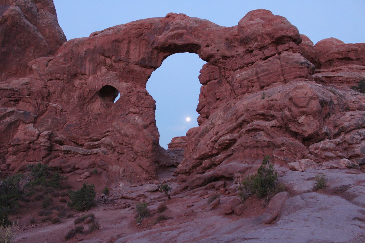Moon seen in the middle of  a red arch formation in the desert.