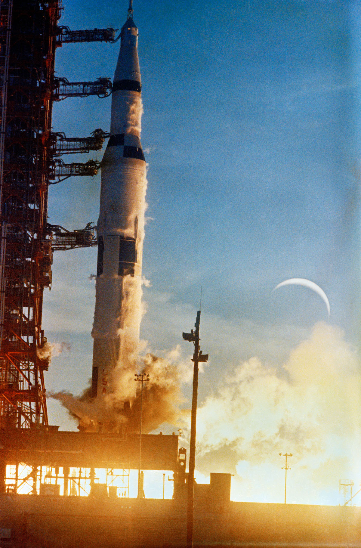 Saturn V rocket launches with great flames, with the Moon in the sky in the background