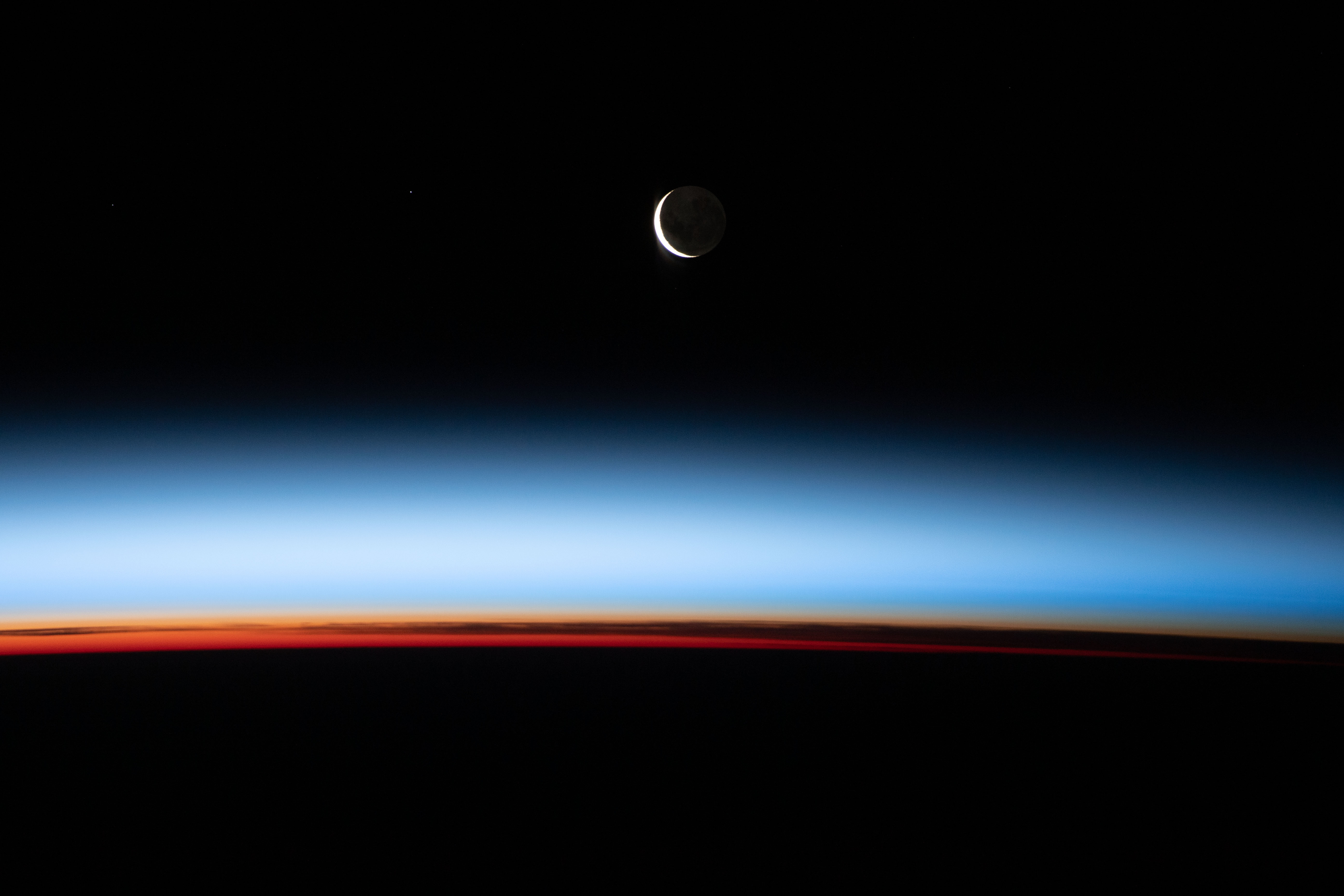 Photograph of a crescent Moon over a rainbow curved atmosphere. 
